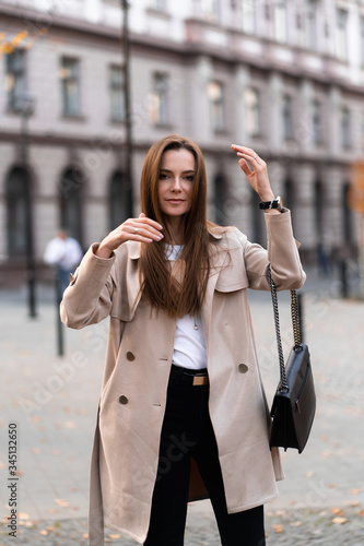 Outdoor photo of brunette lady walking on street background in autumn day.Fashion street style portrait. wearing dark casual trousers, white sweater and creamy coat.Fashion concept.