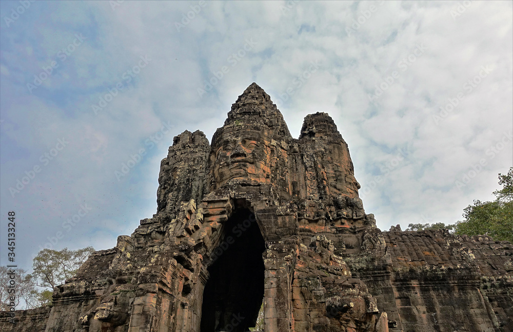 The gates of the ancient Angkor castle in Cambodia against a blue cloudy sky. On the ruins, human faces are carved from stone, looking to all sides of the world.