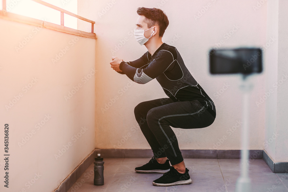 Young healthy sports man in protective mask squatting against phone camera at home. Man fitness instructor in medical face mask recording video tutorial for workout blog. COVID-19, home quarantine Young healthy sports man in protective mask squatting against phone camera at home. Man fitness instructor in medical face mask recording video tutorial for workout blog. COVID-19, home quarantine