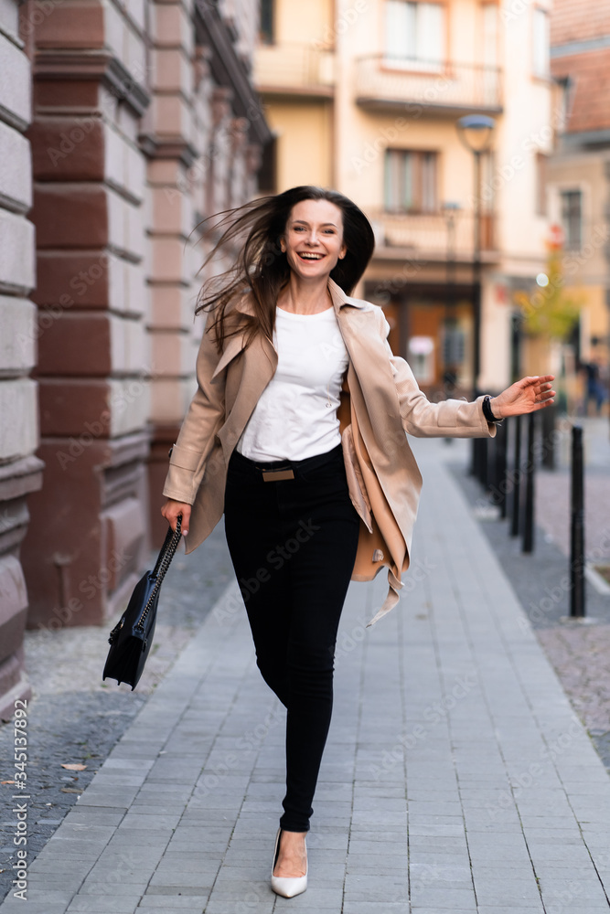 Outdoor photo of brunette lady walking on street background in autumn day.Fashion street style portrait. wearing dark casual trousers, white sweater and creamy coat.Fashion concept.