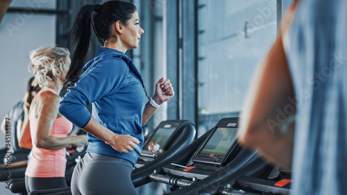 Fit Athletic Woman Running on the Treadmill, Doing Her Fitness Exercise. Muscular Women and Men Actively Training in the Modern Gym. Sports People Workout in Fitness Center. Side View Shot