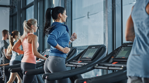 Fit Athletic Woman Running on the Treadmill, Doing Her Fitness Exercise. Muscular Women and Men Actively Training in the Modern Gym. Sports People Workout in Fitness Center. Side View Shot © Gorodenkoff