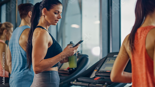 Beautiful Athletic Sports Woman in Gym Running on a Treadmill, Uses Smartphone and Drinks Protein Supplement Hydration Liquid From Tumbler. In Background Fit Athletes Training. Side View