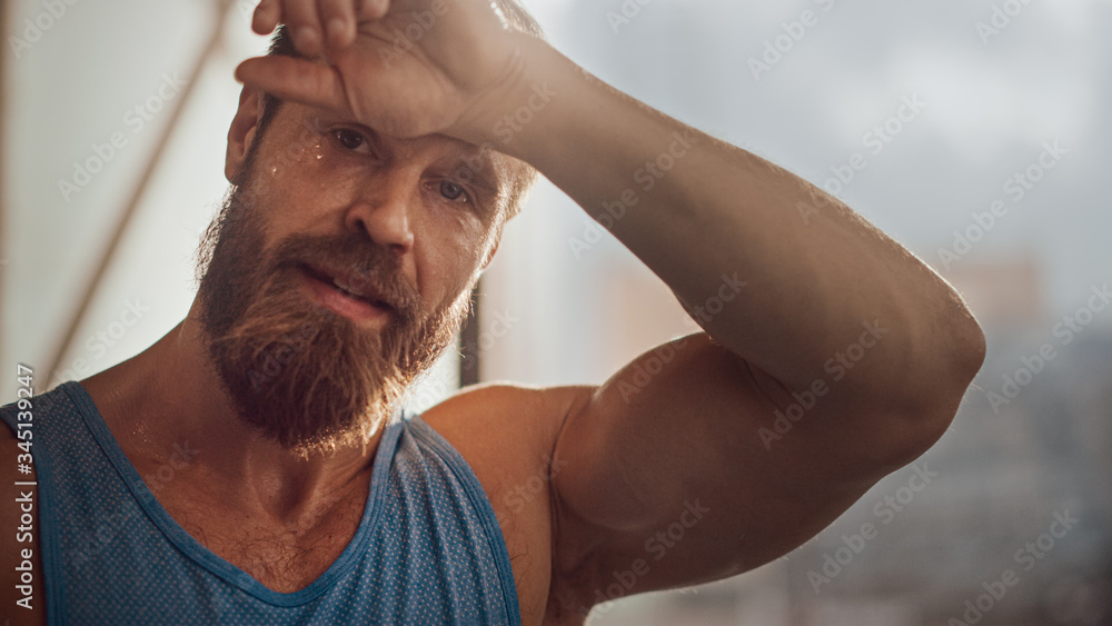 Obraz premium Portrait of Strong Bearded Male Athlete Wearing Sleeveless Shirt Wipes Sweat From His Forehead with His Muscular Hand. Handsome Man after Hardcore Exercise and Training. Man Gets Job Done