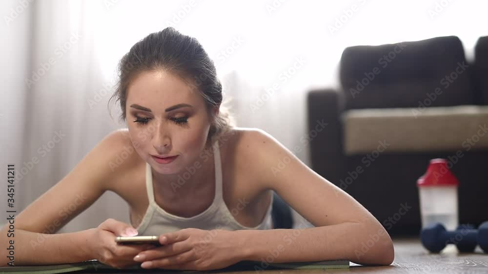 Young woman doing sport exercises at home. She is resting between sets lying on the rug and using a mobile phone. Sports healthy lifestyle. Young woman doing sport exercises at home. She is resting between sets lying on the rug and using a mobile phone. Sports healthy lifestyle.