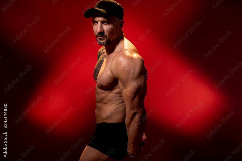 Muscular and strong man in typical bodybuilding pose. Bodybuilder with tattoo in cap on red background. Side view, copy space Muscular and strong man in typical bodybuilding pose. Bodybuilder with tattoo in cap on red background. Side view, copy space