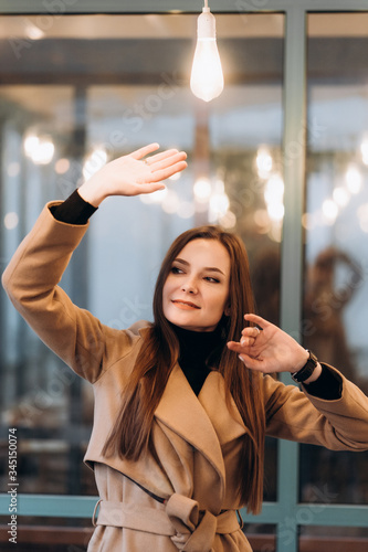 Outdoor fashion portrait of stylish young woman having fun with lamp , laughing, Urban city street style wearing dark casual trousers, white sweater,creamy coat. and sunglasses Fashion concept.