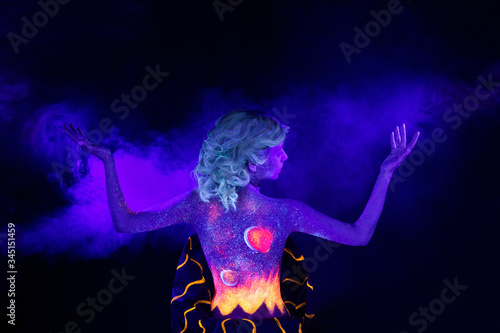 a girl with fluorescent body art under UV lamps in the Studio on a black background with blue smoke stands straight back hands up to the sides