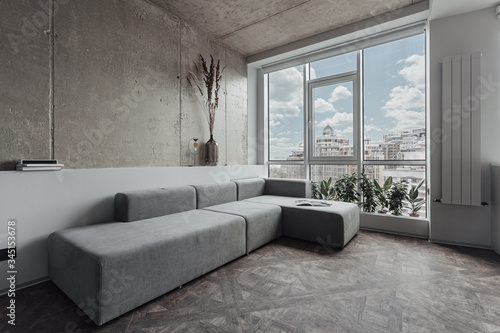 the living room with a long grey sofa near the panoramic window, the walls are decorated as a concrete surface. Minimalist decor, room plants by the window