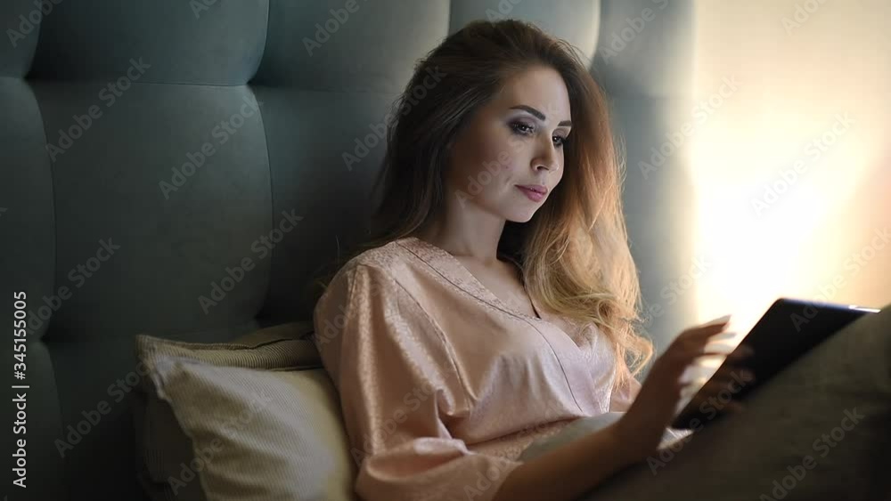 A young woman lying in bed and looking at the good news and photos from a trip on a tablet pc. Girl is reading an e-book in the bedroom. A young woman lying in bed and looking at the good news and photos from a trip on a tablet pc. Girl is reading an e-book in the bedroom.