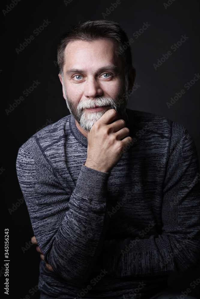 Portrait of a middle-aged man, he stands and looks at the camera, holding by the hand a beard on a dark background Portrait of a middle-aged man, he stands and looks at the camera, holding by the hand a beard on a dark background