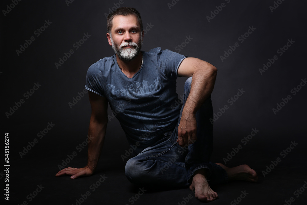 A middle-aged man, sitting full-length, looking at the camera, leg bent at the knee, hand on the knee. On dark background