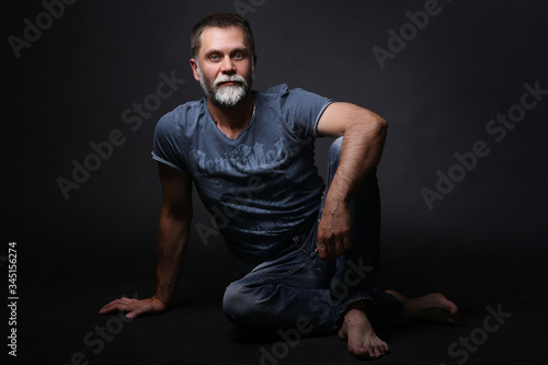A middle-aged man, sitting full-length, looking at the camera, leg bent at the knee, hand on the knee. On dark background © daryakomarova