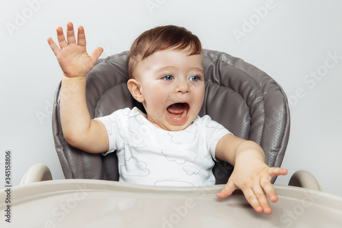 Happy excited adorable baby boy play in high chair with hands up.