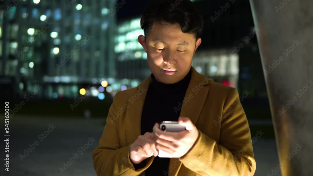 A young Asian man works on a smartphone in a park in a city at night - closeup A young Asian man works on a smartphone in a park in a city at night - closeup