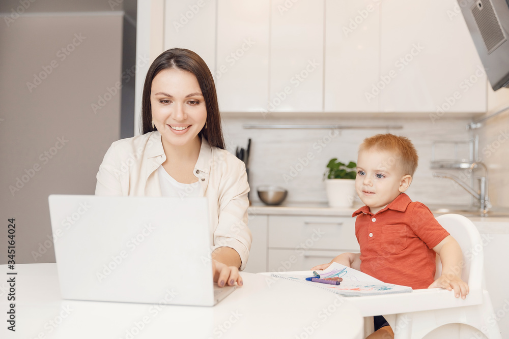 Woman on maternity leave working home online with laptop in kitchen with small child. Concept mom work while in quarantine isolation during Covid-19 Woman on maternity leave working home online with laptop in kitchen with small child. Concept mom work while in quarantine isolation during Covid-19