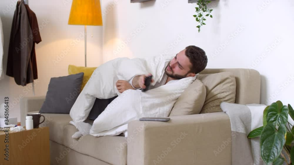 Medium shot of very sick man wrapped in blanket sitting on couch and coughing into paper handkerchief while watching TV at home Medium shot of very sick man wrapped in blanket sitting on couch and coughing into paper handkerchief while watching TV at home