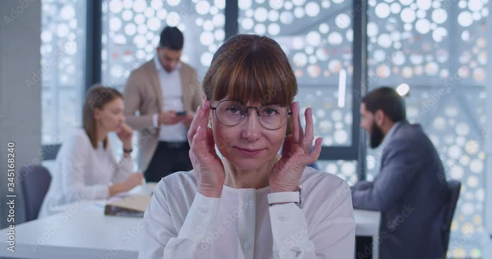 Portrait of middle-aged Caucasian female office worker looking and smiling to camera. Business meeting on background. Senior woman company employee taking on glasses. Portrait of middle-aged Caucasian female office worker looking and smiling to camera. Business meeting on background. Senior woman company employee taking on glasses.