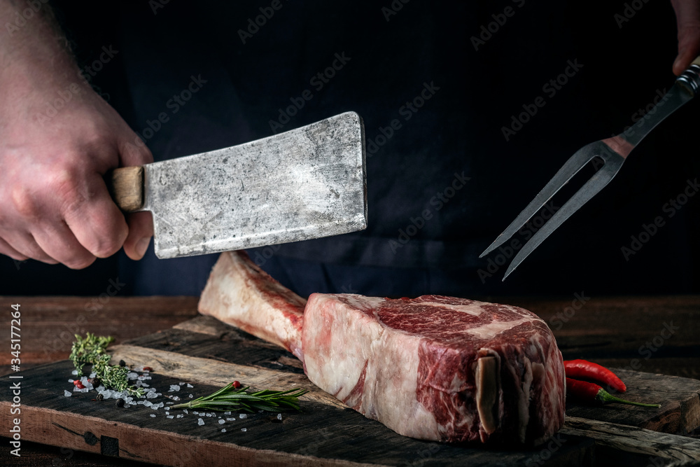 A butcher cuts beef Tomahawk steak on a bone with a cleaver on a wooden cutting Board on a dark background