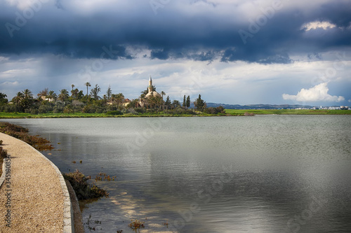 View of Hala Sultan Tekke or Mosque of Umm Haram on the west bank of Salt Lake in Larnaca, Cyprus. Blue dark clouds. Rainy weather. Path (left) along the lake