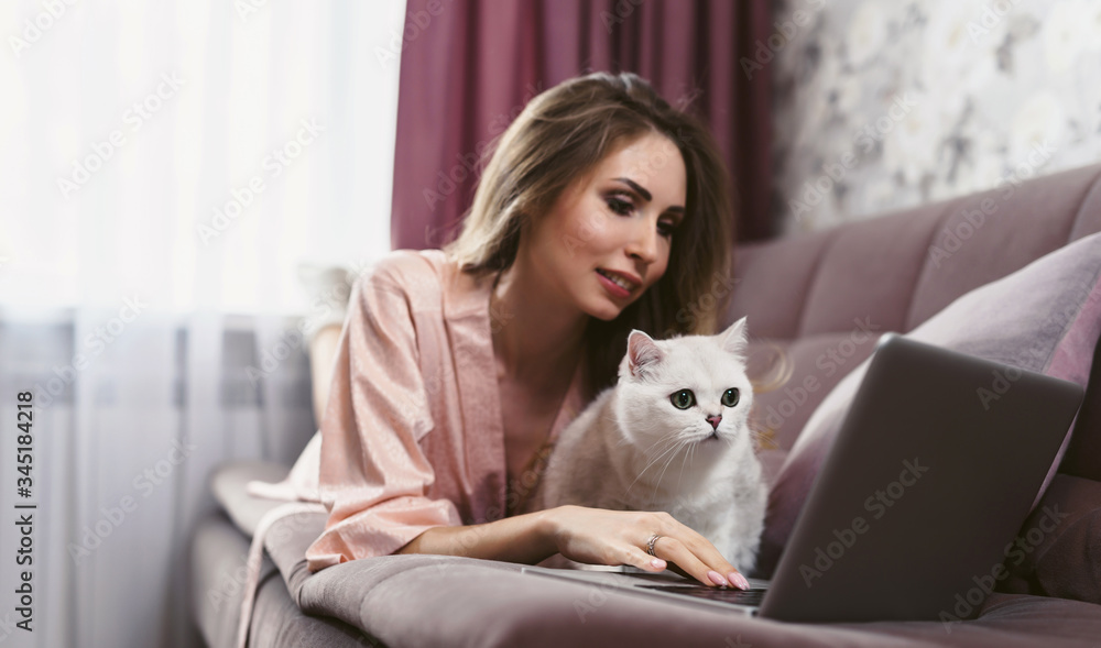 A young pretty woman with a cute cat lies on a sofa and uses a laptop. The pet makes it difficult to work. Pestering cat. Self-isolation. A young pretty woman with a cute cat lies on a sofa and uses a laptop. The pet makes it difficult to work. Pestering cat. Self-isolation.
