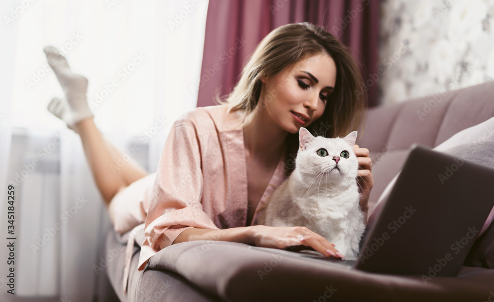 A young pretty woman with a cute cat lies on a sofa and uses a laptop. The pet makes it difficult to work. Pestering cat. Self-isolation. A young pretty woman with a cute cat lies on a sofa and uses a laptop. The pet makes it difficult to work. Pestering cat. Self-isolation.