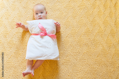 Funny baby girl in a white dress on a pillow with a pink belt from a ribbon on a yellow background for crazy Pillow Challenge due to stay home isolation. Caronavirus Quarantine. photo