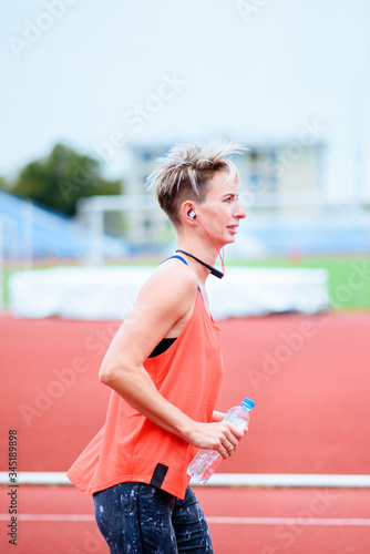 young woman runs in the stadium listening to music and smiling. Holding a bottle of water