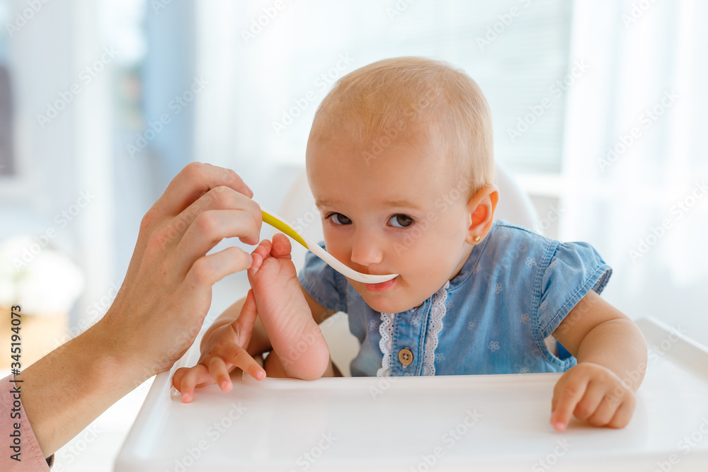 Very cute and funny baby with the spoon in hands eating kids nutrition - fruit puree. Smiling little girl eating in fun pose on white feeding table. Concept of first organic child food and family care Very cute and funny baby with the spoon in hands eating kids nutrition - fruit puree. Smiling little girl eating in fun pose on white feeding table. Concept of first organic child food and family care