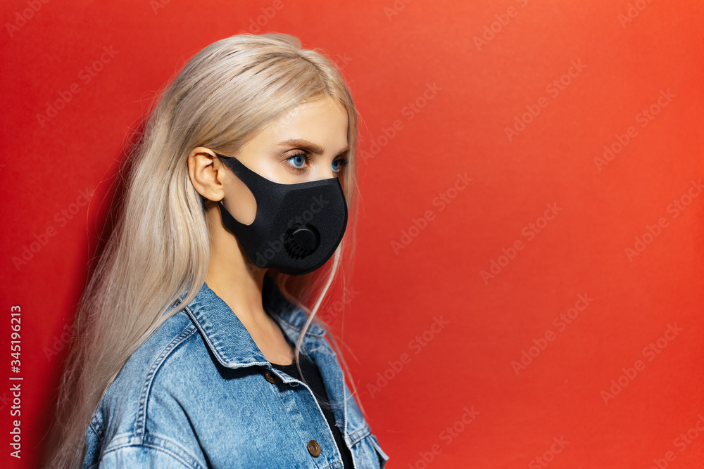 Studio profile portrait of young blonde girl, wearing respiratory face mask of black color, against coronavirus. Background of red color with copy space.