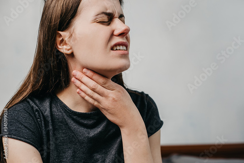 Close up of unhealthy young woman touch neck hard to swallow having discomfort or painful feeling sitting on bed at home, unwell sad girl suffering from angina, sore throat, experience loss of voice photo