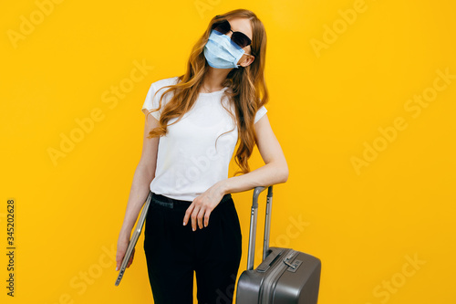 business woman with a protective medical mask on her face, with a laptop and a suitcase on a yellow background. The concept of travel, quarantine, and coronavirus