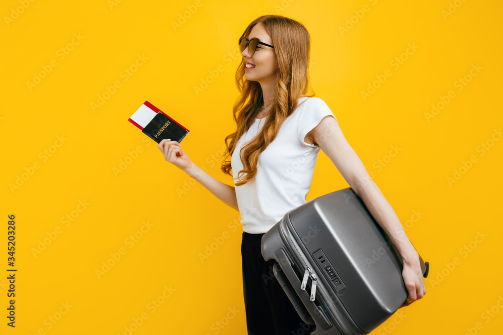 Smiling female passenger in sunglasses, standing with a suitcase and passport with tickets, on a yellow background. Travel concept Smiling female passenger in sunglasses, standing with a suitcase and passport with tickets, on a yellow background. Travel concept