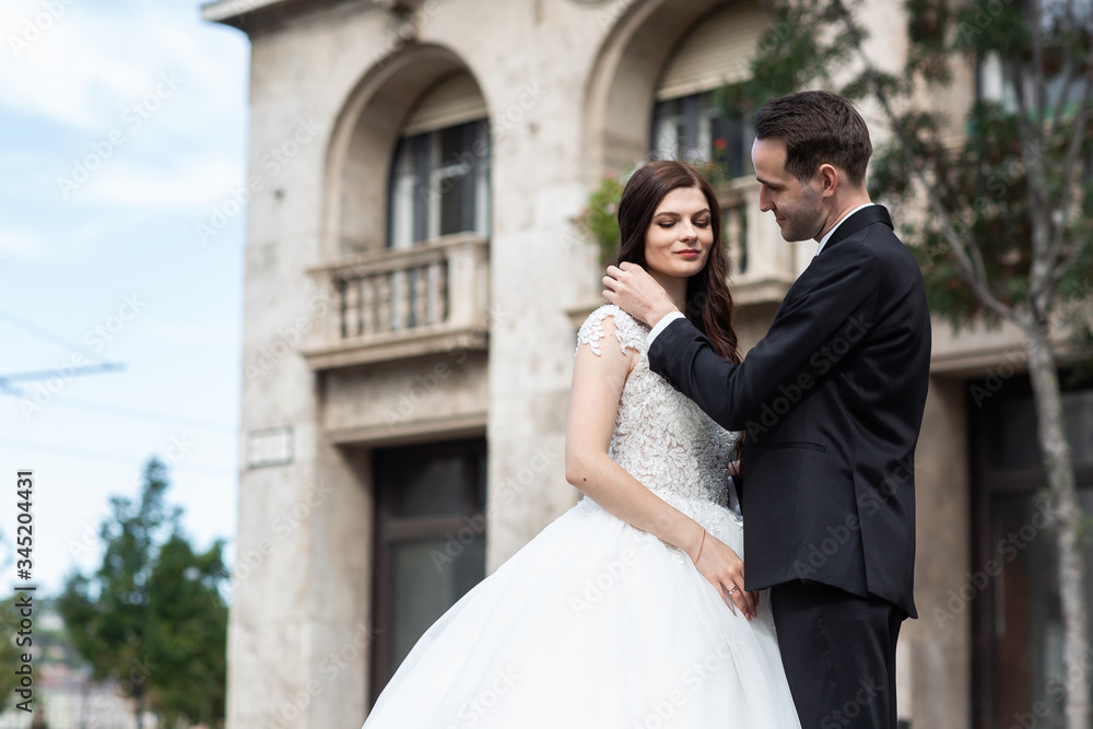 Fototapeta premium Bride and groom hugging in the old town street. Wedding couple walks in Budapest near Parliament House. Caucasian happy romantic young couple celebrating their marriage. Wedding and love concept.
