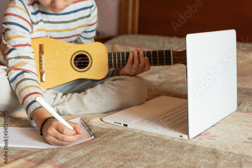 The boy try to learning the timple lesson at home from the internet with his laptop. He writes a new song with chords in his notebook photo