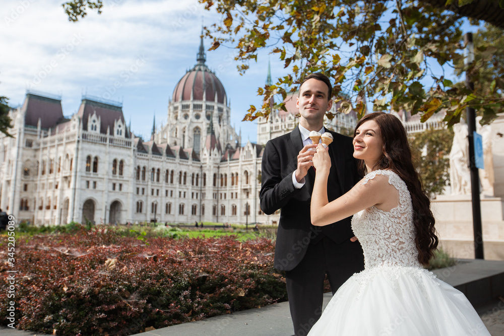 Bride and groom have fun and eating ice cream. Wedding couple sit in Budapest near Parliament House. Caucasian happy romantic young couple celebrating their marriage. Wedding and love concept.