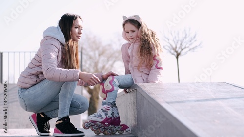 A pretty mother is spending time with her cute daughter. The family is going out for a walk. The kid is going roller-skating. The woman is helping the child to put roller skates on.