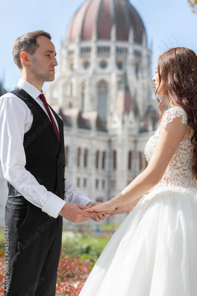 Bride and groom hugging in the old town street. Wedding couple walks in Budapest near Parliament House. Caucasian happy romantic young couple celebrating their marriage. Wedding and love concept. Bride and groom hugging in the old town street. Wedding couple walks in Budapest near Parliament House. Caucasian happy romantic young couple celebrating their marriage. Wedding and love concept.