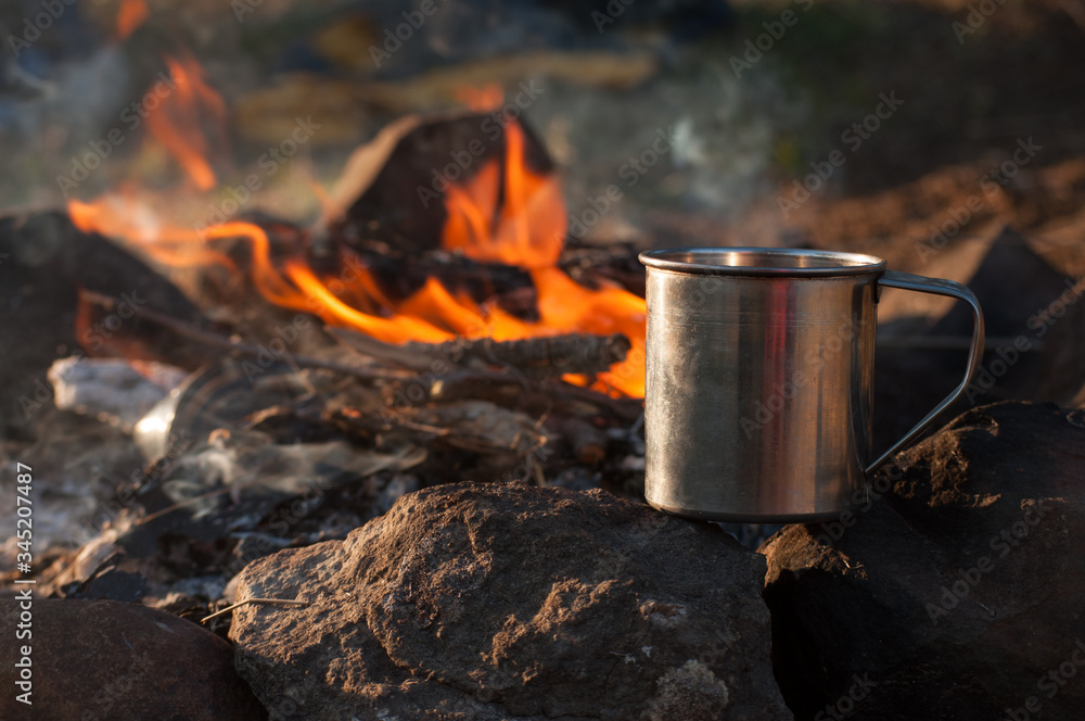 Metal camping mug with hot tea by the fire in the forest. Trekking, camping dishes, bonfire, evening Metal camping mug with hot tea by the fire in the forest. Trekking, camping dishes, bonfire, evening