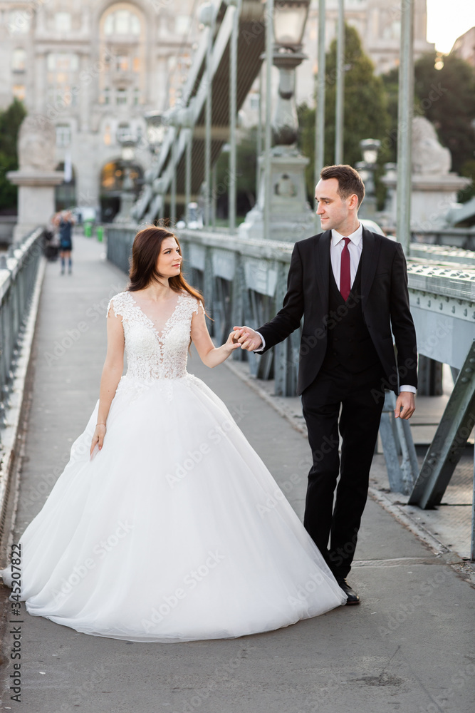 Bride and groom hugging in the old town street. Wedding couple walking on Szechenyi Chain Bridge, Hungary. Happy romantic young couple celebrating their marriage. Wedding and love concept.