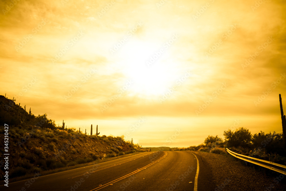 A brilliant sunrise over a road on a hilltop with saguaro cacti on the sides of the road in Arizona.