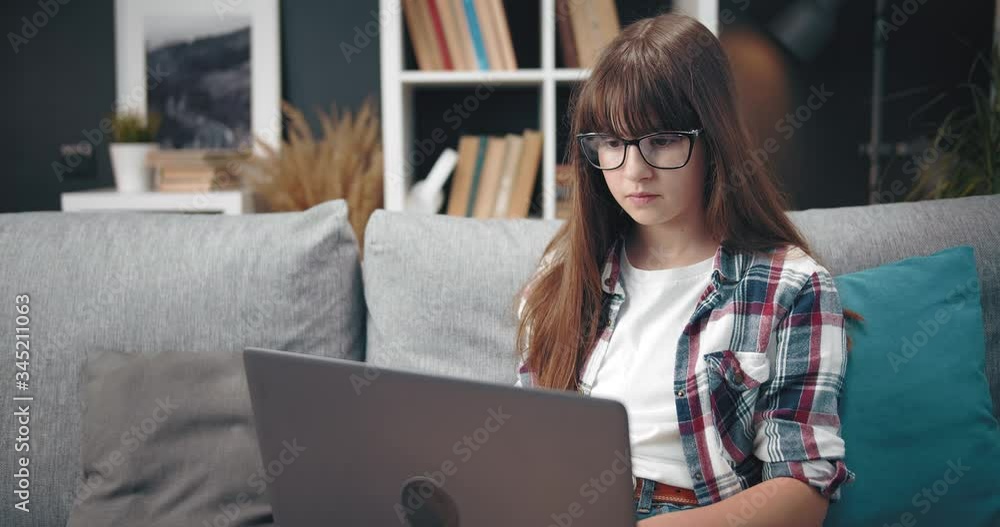 Beautiful girl with dark hair using portable laptop for studying while sitting on grey couch. Cute teenager in domestic outfit spending free time for education. Beautiful girl with dark hair using portable laptop for studying while sitting on grey couch. Cute teenager in domestic outfit spending free time for education.