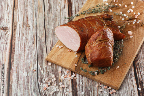 Smoked ham on a chopping board. Sliced smoked gammon on a wooden table with addition of fresh herbs and aromatic spices. Natural product from organic farm, produced by traditional methods