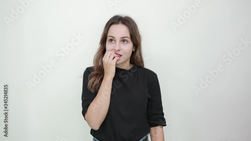 Cute Young Brunette Woman In Black T-Shirt, Blue Jeans With Belt On Gray Background, Confused Girl Does Not Know How To React To The Situation And Bites Nails, Holds Hands Near Mouth