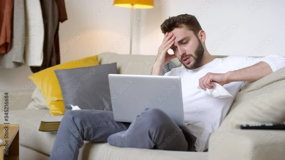Tracking shot of very sick man with headache and cough sitting on couch and talking on video call with someone using laptop Tracking shot of very sick man with headache and cough sitting on couch and talking on video call with someone using laptop