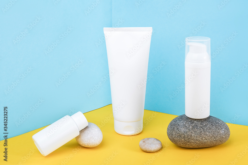 White blank cosmetics bottles, tube of cream, lotion for body, face, hand or other cosmetic product and gray rocks on blue yellow background. Mockup Front view, Corner composition Copy space White blank cosmetics bottles, tube of cream, lotion for body, face, hand or other cosmetic product and gray rocks on blue yellow background. Mockup Front view, Corner composition Copy space
