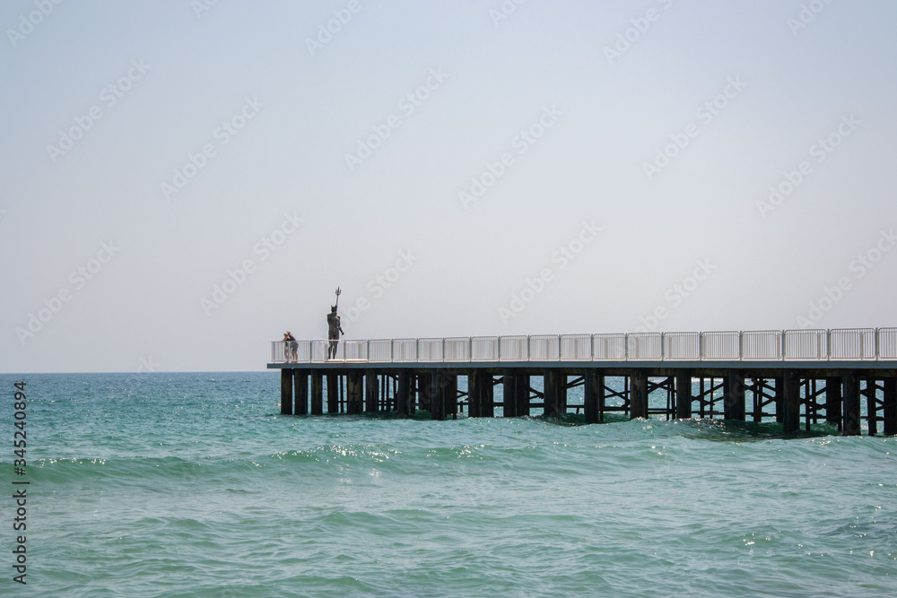 People walking on a pier with statue of Poseidon, bridge near the beach, blue sea and sky