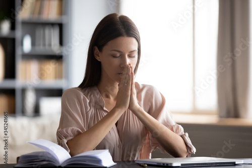 Calm woman with closed eyes putting hands in prayer, sitting at desk at home, wishful young female making wish, difficult decision, asking help, thinking about difficult work, problem solution