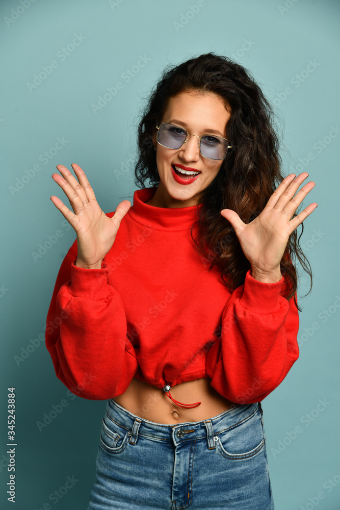 Portrait of smiling adult slim woman with long curly hair in red top and blue jeans holding hands up with open palms Portrait of smiling adult slim woman with long curly hair in red top and blue jeans holding hands up with open palms