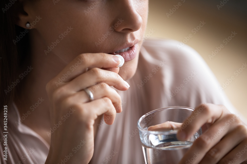 Close up young woman taking white round pill, holding water glass, unhealthy female taking painkiller to relieve headache, antidepressant or antibiotic medicine, emergency treatment Close up young woman taking white round pill, holding water glass, unhealthy female taking painkiller to relieve headache, antidepressant or antibiotic medicine, emergency treatment